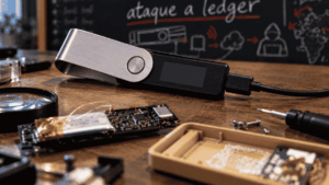 Primer plano: hardware wallet Ledger. Fondo: pizarra donde se lee "ataque a ledger"