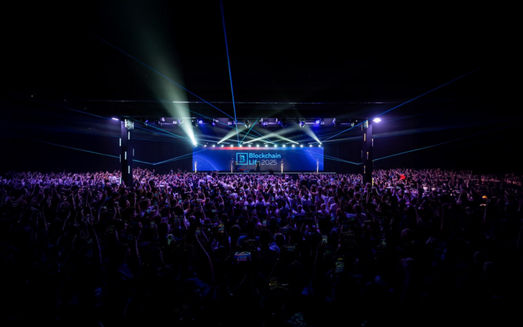 Sala llena de asistentes frente al escenario principal de Blockchain Life 2025, con luces láser y pantalla del evento al fondo.