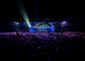 Sala llena de asistentes frente al escenario principal de Blockchain Life 2025, con luces láser y pantalla del evento al fondo.