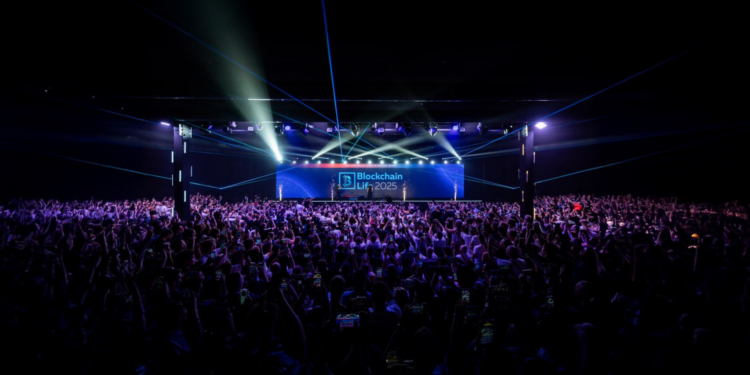Sala llena de asistentes frente al escenario principal de Blockchain Life 2025, con luces láser y pantalla del evento al fondo.