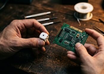 Close-up of two human hands in warm side light, one hand holding a single white precision die, the other holding a bare green circuit board with visible chips and solder points, dark wooden workbench, shallow depth of field