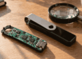 Sobre una mesa: dispositivo Ledger abierto, dispositivo Ledger cerrado y una lupa.